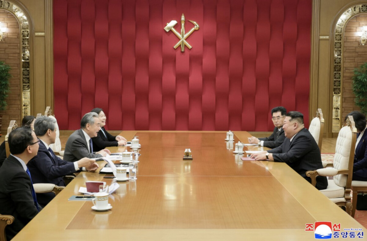 diplomatic meeting in a wood floored conference room with officials seated across a long table a red padded wall behind them and a gold emblem above